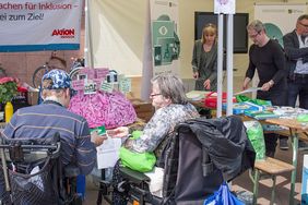 Zwei Rolli-Fahrer sitzen vor einem Gehirn aus Pappmach&eacute; an einem Stand. Im Hintergrund ist der ehemalige Behindertenbeauftragte von Sachsen, Stephan P&ouml;hler, zu sehen.