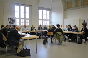 Menschen sitzen in U-Form am Tisch, in der Mitte sitzen Gebärdensprachdolmetscher