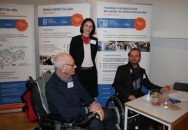 drei Personen: eine &auml;ltere Person im Rollstuhl mit grauem Haar von der Seite, in der Mitte steht eine Frau mit halblangen Haaren und Brille, rechts ist ein weiterer Mann in Rollstuhl, sie stehen hinter einem Stand. Im Hintergrund sind Rollups zum Projekt.