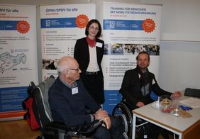 drei Personen: eine &auml;ltere Person im Rollstuhl mit grauem Haar von der Seite, in der Mitte steht eine Frau mit halblangen Haaren und Brille, rechts ist ein weiterer Mann in Rollstuhl, sie stehen hinter einem Stand. Im Hintergrund sind Rollups zum Projekt.
