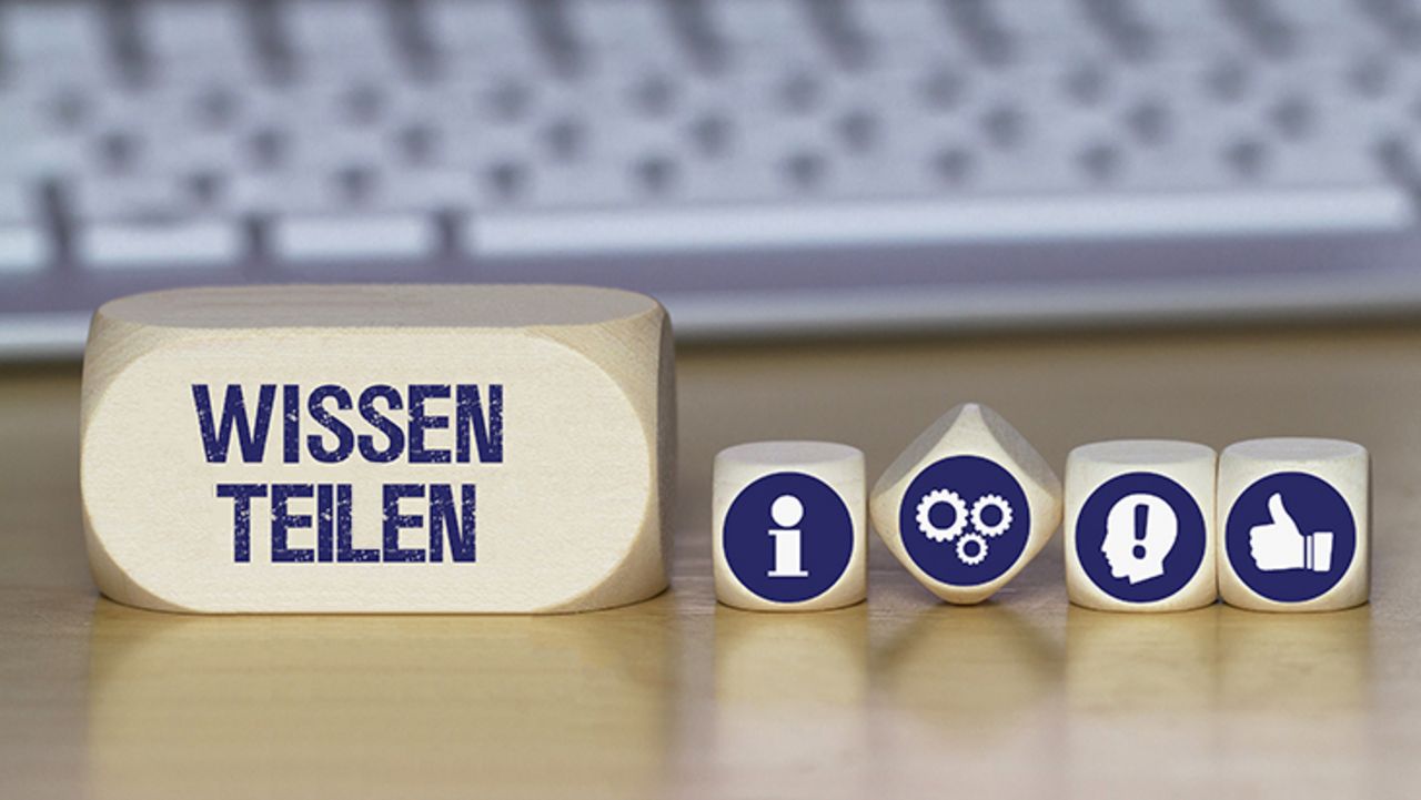 Foto von Holzwürfeln vor einer Tastatur. Auf einem Würfel steht "Wissen teilen". Auf den anderen sind Symbole zu sehen: i für Information, Zahnräder, ein Kopf mit einem Ausrufezeichen darin und ein nach oben gerichteter Daumen