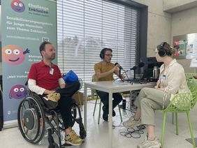 Thomas Naumann sitzt im Rollstuhl an einem Tisch. Auf dem Tisch stehen zwei Mikrofone, an denen Taddeus Helm und eine junge Frau sitzen und Kopfhörer tragen.