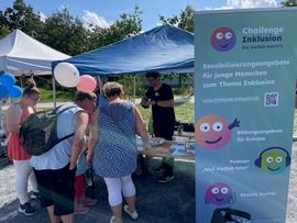  Drei Frauen stehen in der Sonne vor dem Stand der LAG SH Sachsen und sehen sich die Materialien an. Daneben steht ein gro&szlig;es buntes Roll-Up, auf dem Informationen zum Projekt Challenge Inklusion zu lesen sind. 