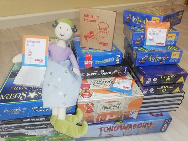 Glücksfee als Kuscheltier sitzt auf mehreren Stapeln aus Spielen, Büchern und hält eine Tüte von Aktion Mensch Inklusion blüht in der Hand