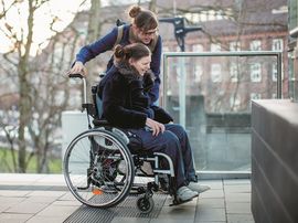 Frau im Rollstuhl wird durch Assistentin begleitet