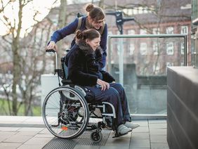 Frau im Rollstuhl wird durch Assistentin begleitet