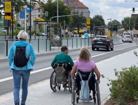 Zwei Rollstuhlfahrer fahren auf einem B&uuml;rgersteig entlang. Eine Frau l&auml;uft neben Ihnen