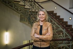 Verena Bentle auf dem Foto, sie ist eine Frau mit blonden mittellangen Jahren und brauner Jacke, steht im Treppenhaus