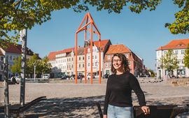 Frau mit schwarzem-lockigem Haar und Brille im schwarzen Shirt steht auf dem Marktplatz von Bischofswerda, Frau l&auml;chelt in die Kamera