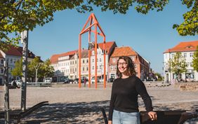 Frau mit schwarzem-lockigem Haar und Brille im schwarzen Shirt steht auf dem Marktplatz von Bischofswerda, Frau lächelt in die Kamera