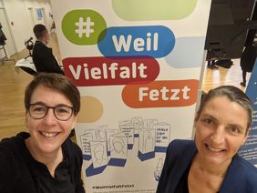 Mitarbeiterinnen des Inklusionsnetzwerk Sachsen Susanne R&ouml;&szlig;ner (links) und Heike Engelien (rechts) vor dem Roll-Up #WeilVielfaltfetzt am Stand des Inklusionsnetzwerk Sachsen