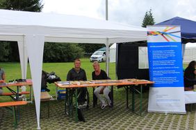 Zwei Mitarbeiter des Inklusionsnetzwerk Sachsen, Falk Friedrich (links) und Angela Werner (rechts) sitzen an einem Tisch am Stand des INWS. Auf dem Tisch liegen Bauteile eines Mobilit&auml;tsbaukastens f&uuml;r Rollstuhlfahrer. Die beiden sitzen unter einem Zeltdach an dem ein Roll-Up des INWS befestigt ist.
