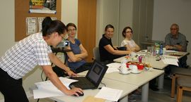 Blick auf Gespr&auml;chsrunde - Frau erkl&auml;rt Teilnehmern eine Situation und tippt dabei auf dem Laptop