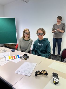 Zwei Frauen sitzen an einem Tisch und gucken in die Kamera. Sie tragen Simulationsbrillen. Hinter ihnen steht eine Mitarbeiterin des Jobcenters. Auf dem Tisch liegen Aufgabenzettel und weitere Brillen.