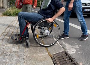 Das Foto zeigt in einer totalen Ansicht, wie ein Rollstuhlnutzer den &Uuml;bergang von der Stra&szlig;e zu einem Gehweg &uuml;berwindet. Dabei wird ihm von einer weiteren Person geholfen, indem sie von hinten schiebt. Das &uuml;berwinden der Schwelle erscheint m&uuml;hevoll.