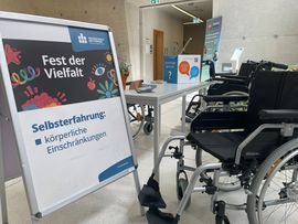 Auf einem Aufsteller steht "Fest der Vielfalt - Selbsterfahrung k&ouml;rperliche Behinderungen". Daneben stehen zwei leere Rollst&uuml;hle an einem Tisch voller Flyer.