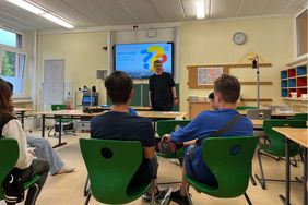 Ein Mann referiert vor Schüler*innen in einem Klassenraum. Er hat einen Bart, ist Ende 30, hat eine Brille und trägt schwarze Kleidung. Drei Schüler, ein Mädchen und 2 Jungen, sind von hinten zu sehen während sie an ihren Schultischen sitzen. An der Tafel ist eine Powerpoint Präsentation zum Thema Inklusion zu sehen.