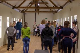 zwei Personen erz&auml;hlen zu einem Exponat in der Gedenkst&auml;tte Pirna-Sonnenstein, darum stehen Frauen und M&auml;nner und h&ouml;ren zu