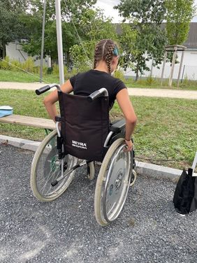Ein Mädchen sitzt in einem Rollstuhl und versucht, über eine Stufe auf eine Wiese zu gelangen. Sie trägt ein schwarzes T-Shirt und ihre langen Haare sind in zwei französischen Zöpfen zusammengebunden.