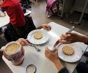 Tisch mit Geschirr, Brot und Frischk&auml;se. Zwei Kinder versuchen mit verbundenen Augen das Brot zu schmieren. 