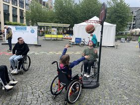 Ein junges Mädchen sitzt im Rollstuhl und wirft einen Korb beim Rollstuhl-Basketball. Zwei junge Männer Anfang Zwanzig sitzen im Rollstuhl daneben. Alle sehen sehr erfreut aus und haben Spaß.