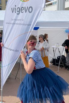 Ein weiblicher Clown lehnt sich an die Beachflag von Vigevo und grinst in die Kamera.