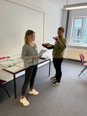 Lara Hanske steht vor den Teilnehmer*innen und liest etwas von einem Zettel ab. Sie tr&auml;gt ihre halblangen blonden Haare in einem Zopf und hat eine schwarze Hose und ein graues Oberteil an. Stephan Zapke h&auml;lt verschiedene Simulationsbrillen in der Hand. Er ist sichtbar sehbehindert und tr&auml;gt eine schwarze Hose und einen gr&uuml;nen Pullover. Er hat kurze dunkelblonde Haare.