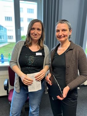 Kerstin Helm und Heike Engelien l&auml;cheln in die Kamera. Kerstin Helm hat halblange braune Haare, tr&auml;gt eine graue Strickjacke, ein schwarzes Tshirt und Jeans. Heike Engelien tr&auml;gt ihre dunklen Haare im Zopf und hat eine schwarze Hose, ein schwarzes Tshirt und eine braune Strickjacke an. Beide halten Zettel mit Quizaufgaben in der Hand. 