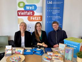 [Translate to Leichte Sprache:] Das Team der LAG SH Sachsen sitzt am Stand der LAG SH Sachsen auf dem Bildungskongress in G&ouml;rlitz. Katja R&ouml;&szlig;ner (links), Kerstin Helm (mitte) und Stephan Zapke (rechts). Auf dem Tisch vor dem Team liegen verschiedene Materialen aus den Projekten der LAG SH Sachsen aus.