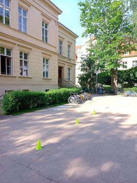 [Translate to Leichte Sprache:] Der Schulhof der Heise Schule in G&ouml;rlitz. Auf ndem Schulhof ist ein Rollstuhlparcour mit gelben Kegeln abgesteckt. Vor dem Schulgeb&auml;ude steht eine Hecke, vor der 4 leere Rollst&uuml;hle stehen