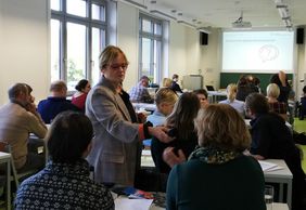 Annegret Haage h&auml;lt ein Mikrofon in Richtung eines Teilnehmers und steht mitten zwischen den Tischen mit Teilnehmenden