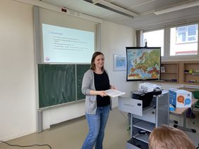 [Translate to Leichte Sprache:] Zu sehen ist Kerstin Helm, die vor einer Tafel steht, w&auml;hrend sie einen Vortrag h&auml;lt. Sie hat braune Haare, l&auml;chelt, tr&auml;gt eine Jeans, ein schwarzes Oberteil und eine graue Strickjacke. Im Hintergrund sieht man eine Schultafel mit einer PowerPoint Pr&auml;sentation. Rechts im Bild steht eine Landkarte von Europa f&uuml;r den Geographieunterricht und ein Tisch mit technischen Ger&auml;ten.