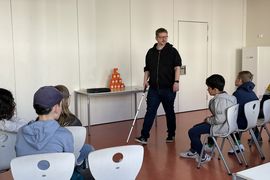 Der Dozent zeigt einer Grundschulklasse, wie man einen Blindenlangstock richtig verwendet. Er tr&auml;gt einen schwarzen Kaputzenpullover und eine schwarze Jeans. In der rechten Hand h&auml;lt er den Blindenlangstock, w&auml;hrend er zur Klasse spricht. Die Kinder schauen interessiert zu.