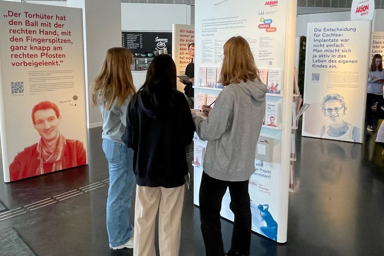 Drei Mädchen stehen vor eine Stele der Wanderausstellung "Weil Vielfalt fetzt" und lesen sich die Texte durch. Im Hintergrund sieht man weitere Schüler*innen und Stelen der Ausstellung. Die Stele auf der linken Seite zeigt Florian Eib. Er ist ein junger Mann mit kurzen dunklen Haaren und einem Tuch um den Hals, der freundlich in die Kamera lächelt. Das Foto wurde rot eingefärbt. Daneben steht folgender Text: "Der Torwart hat den Ball mit der rechten Hand, mit den Fingerspitzen, ganz knapp am rechten Pfosten vorbeigelenkt."