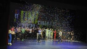 Viele junge Menschen stehen auf einer Theaterbühne. Die Scheinwerfer leuchten hell und hinter der Gruppe steht sehr groß Unart an der Wand. Einige tragen Kostüme, andere haben sich einfach schick gemacht. Einzelheiten lassen sich schlecht erkennen. Die Jugendlichen stehen in einem bunten Konfettiregen. Er regnet von oben auf Bühne herab. Alle freuen sich. Viele von ihnen klatschen in die Hände. Es ist der Abschluss des Unart-Finale 2022 im Kleinen Haus.