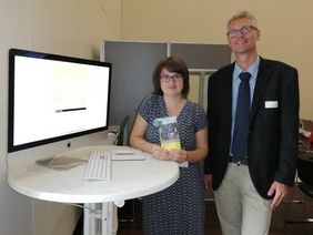 Frau mit dunklen Haaren und gro&szlig;er Mann stehen nebeneinander an einem Rechner und blicken frontal